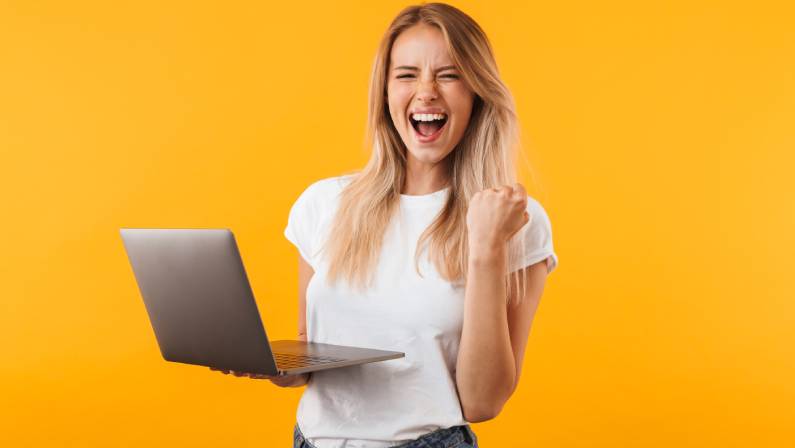 Excited young blonde girl holding laptop and celebrating success of booking a rental car early.